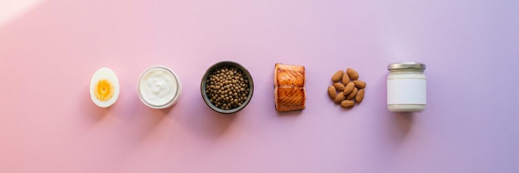 Alimentos proteicos cotidianos alineados sobre un fondo pastel, entre ellos huevo, yogur, lentejas, salmón, almendras y una crema proteica