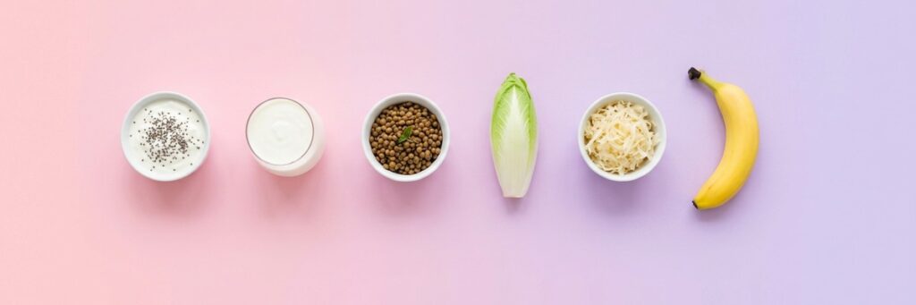 Alimentos para el confort digestivo: yogur con chía, kéfir, lentejas, achicoria, col fermentada y plátano sobre fondo pastel