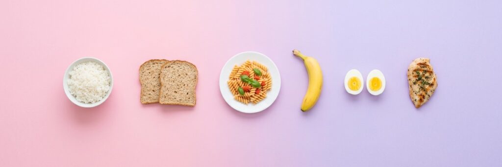 Menos proteína, menos peso: el ensayo isocalórico que obliga a matizar 5 Alimentos con menos proteína y más hidratos sobre fondo pastel: arroz, pan, pasta, plátano, huevo y pollo
