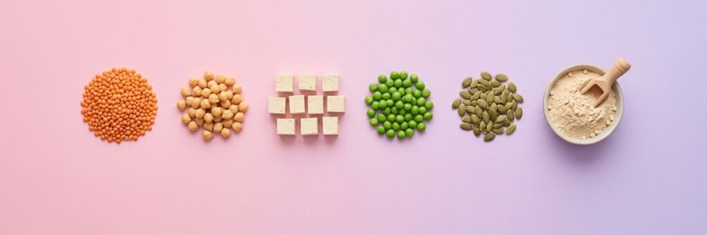 Veganos activos con baja proteína: misma recuperación, más agujetas 2 Fila horizontal de lentejas rojas, garbanzos, tofu, guisantes, semillas de calabaza y un cuenco de proteína de guisante sobre fondo degradado rosa y lavanda